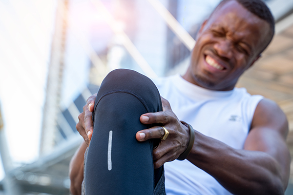 African American male runner bends over clutching his knee while in intense pain from an acute knee injury